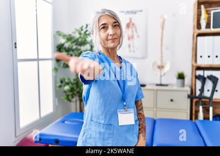Grauhaarige Frau mittleren Alters trägt Physiotherapeutin Uniform in der medizinischen Klinik lächelnd freundlich mit Handschlag als Begrüßung und Begrüßung. Erfolgreich Stockfoto