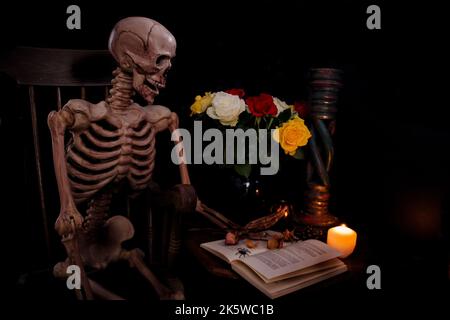 Skelett sitzt an einem Tisch in Schaukelstuhl mit frischen Rosen und toten Rosen ein Buch von Kerzenlicht zu halloween lesen Stockfoto