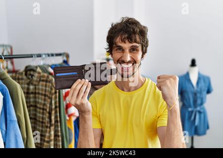 Junger hispanischer Mann, der im Einzelhandelsgeschäft eine Ledergeldbörse hält und stolz schreit und mit erhobenem Arm den Sieg und den Erfolg feiert Stockfoto
