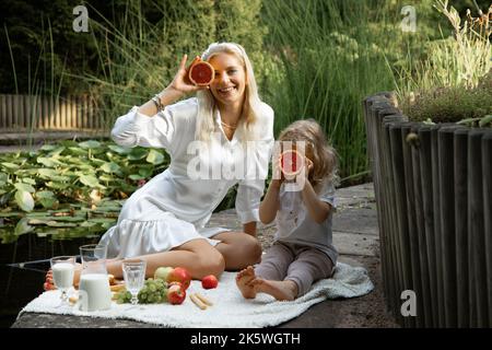 Porträt einer lachenden Familie von junger Mutter und kleiner Tochter, die auf weißem Karli sitzen und Grapefruits in der Nähe des Gesichts halten. Stockfoto