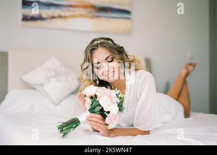 Die Braut im weißen Bademantel liegt auf dem Bauch auf dem Bett und blickt auf einen Blumenstrauß Stockfoto