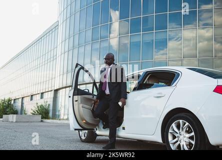 Porträt eines erfolgreichen afroamerikanischen Geschäftsmannes in einem Anzug, der vor dem Hintergrund eines Glasgebäudes aus seinem Privatwagen aussteigen konnte Stockfoto