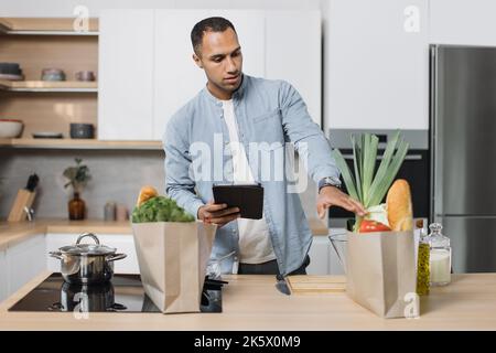 Attraktiver junger araber, der sich ein Online-Rezept auf einem digitalen Tablet ansieht. Schöner arabischer Mann, der im Internet nach Rezepten surft, während er in seiner modernen Küche vegetarischen Salat zubereitet. Stockfoto
