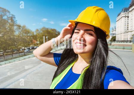 Porträt einer jungen Ingenieurin, die lächelt, mit gelbem Helm, grüner Weste und blauer Kleidung, die mit ihrem Handy vor dem Stern ein Selfie im Freien macht Stockfoto