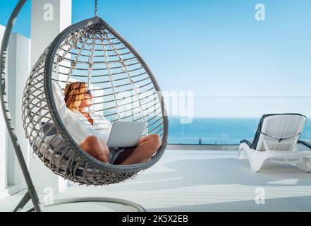 Schöne Frau mittleren Alters, die in einem bequemen Hängesessel auf der offenen Terrasse mit einem modernen Laptop sitzt und die Sonnenstrahlen des Sonnenuntergangs genießt. Mo Stockfoto
