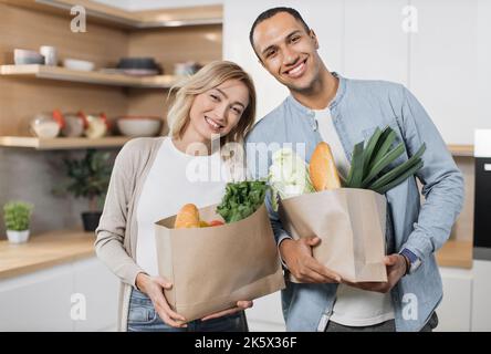 Fröhlicher, junger indischer Mann und Frau stehen zusammen in der Küche, lächeln, blicken auf die Kamera und packen die Lebensmittelpapiertasche aus. Konzept der Familie, häusliche Lebensweise und gesunde Ernährung. Stockfoto