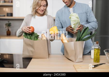 Beschnittenes Bild eines glücklichen jungen Ehepaares, das vom Einkaufen nach Hause kommt und zusammen Papiertüten mit Lebensmitteln in der Küche auspackt. Haushalts- und Beziehungskonzept. Stockfoto