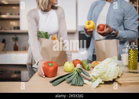 Beschnittenes Bild eines glücklichen jungen Ehepaares, das vom Einkaufen nach Hause kommt und zusammen Papiertüten mit Lebensmitteln in der Küche auspackt. Haushalts- und Beziehungskonzept. Stockfoto