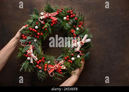 Weihnachtskranz aus natürlichen immergrünen Zweigen mit roten Stechbeeren in Frauenhand auf braunem Holzhintergrund. Festliche Dekoration für Weihnachten. Stockfoto