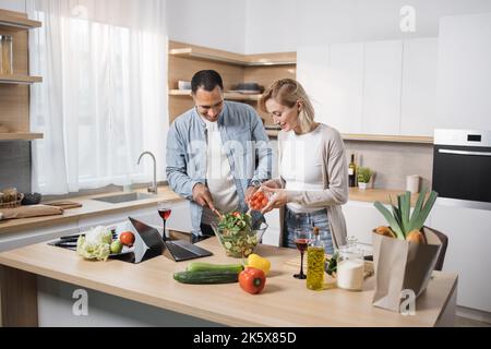 Junge attraktive Paar in der Liebe Vorbereitung Salat aus frischem Gemüse. Ein hübscher, sportlicher Mann, der Scheiben von Zutaten mischt, und eine blonde, charmante Frau, die Olivenöl in eine Glasschale gießt. Stockfoto