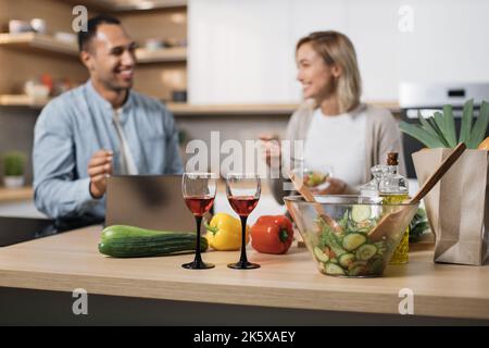 Konzentrieren Sie sich auf Gläser mit Rotwein. Fröhliches Paar mit Laptop essen Diätsalat aus frischem Bio-Gemüse und sprechen miteinander, lächeln, entspannen nach dem Arbeitstag in der modernen Küche zu Hause. Stockfoto