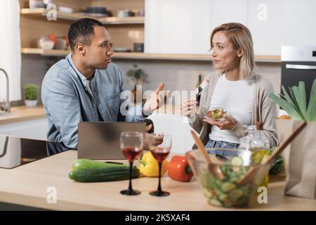 Fröhliches multinationales Paar mit Laptop, das Diätsalat aus frischem Bio-Gemüse isst und mit einander spricht, lächelt und entspannt nach dem Arbeitstag in der modernen Küche zu Hause. Stockfoto