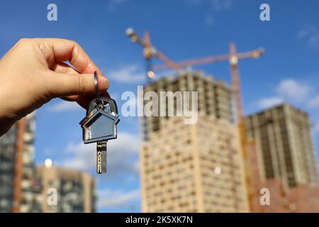 Immobilienmakler, Schlüsselanhänger in Form eines Hauses und Schlüssel in weiblicher Hand auf dem Hintergrund von Baukräne und Neubauten. Byung Wohnung oder Miete Stockfoto