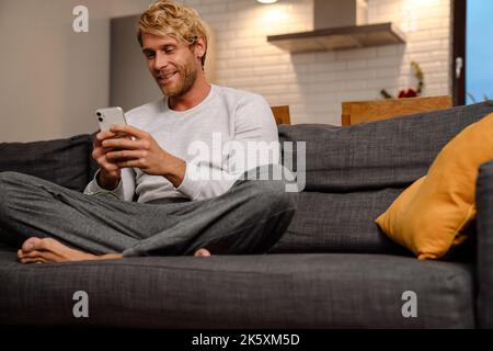 Blonde bärtige Mann mit Handy, während auf dem Sofa zu Hause sitzen Stockfoto