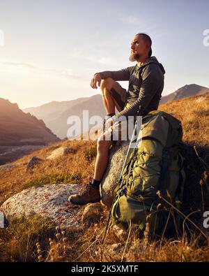 Portrait des Bartwanderers Tourist mit großem Rucksack und Trekkingstöcken, die auf dem Felsen sitzen, gegen den wunderschönen Sonnenuntergang im Bergtal. Übertrumpfst Stockfoto