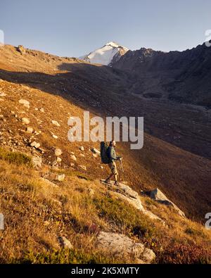 Portrait eines Bartwanderers mit großem Rucksack und Trekkingstöcken, der gegen den wunderschönen Sonnenuntergang im Bergtal den Felsen hinunter läuft. Out Stockfoto