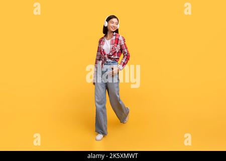 Junge japanische Dame, die Musik in kabellosen Kopfhörern hört, geht über gelben Studiohintergrund, in voller Länge Stockfoto