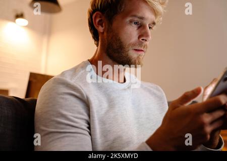 Blonde bärtige Mann mit Handy, während auf dem Sofa zu Hause sitzen Stockfoto