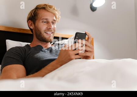 Blonde bärtige Mann mit Handy, während im Bett zu Hause liegen Stockfoto