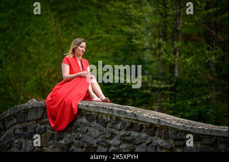 Eine Frau in einem roten Kleid sitzt auf einer alten gotischen Steinmauer, ein Fotoshooting an verlassenen und vergessenen Orten. Stockfoto