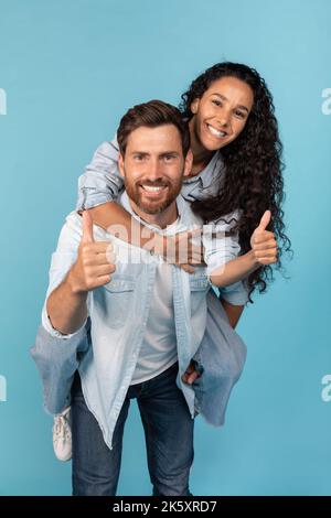 Happy Millennial Caucasian guy Holding arab female on his back, have fun, show thumbs up, pärchen enjoy Freedom Stockfoto