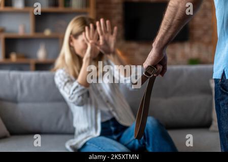 Verzweifelte europäische Frau mittleren Alters, die im Wohnzimmer von einem aggressiven Mann mit Gürtel geschlagen wurde Stockfoto