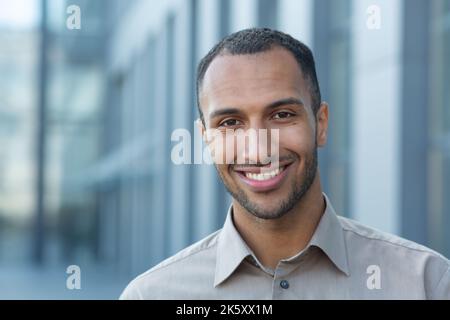 Nahaufnahme eines Portraits eines jungen afroamerikanischen Studenten, lächelnder Mann und Blick auf die Kamera, Geschäftsmann vor dem Bürogebäude mit Hemd. Stockfoto