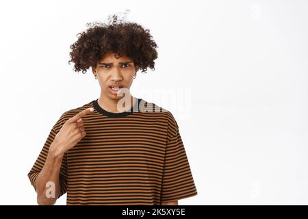 Verwirrt Kerl zeigt auf sich selbst, Blick mit verwirrt schockiert Gesicht, sprechen Sie mit mir, stehen über weißem Hintergrund Stockfoto
