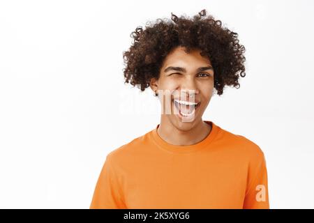 Nahaufnahme eines gutaussehenden hispanischen Mannes mit lockigen Haaren, lächelnd und lachend, frech aussehend, mit orangefarbenem T-Shirt, stehend über weißem Studiohintergrund Stockfoto