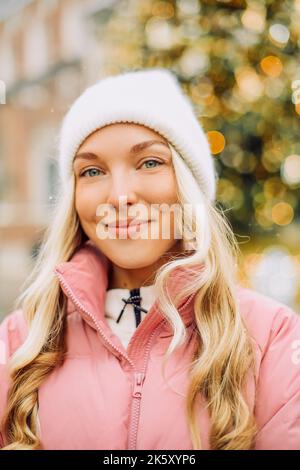 Eine niedliche Blondine in einem Hut und einer rosa Jacke vor dem Hintergrund eines Weihnachtsbaums auf der Straße. Winterspaziergang, Neujahr, Festtag. Stockfoto