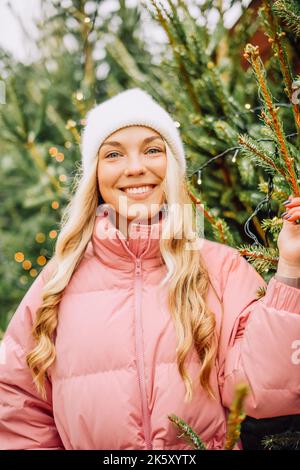 Eine niedliche Blondine wählt einen Weihnachtsbaum für das neue Jahr. Eine Frau in einem Hut und einer rosa Jacke lächelt bei schneebedecktem Wetter auf die Kamera. Winterferien und n Stockfoto