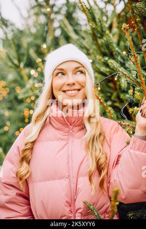 Eine niedliche Blondine wählt einen Weihnachtsbaum für das neue Jahr. Eine Frau in einem Hut und einer rosa Jacke lächelt bei schneebedecktem Wetter auf die Kamera. Winterferien und n Stockfoto