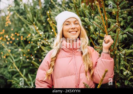 Eine niedliche Blondine wählt einen Weihnachtsbaum für das neue Jahr. Eine Frau in einem Hut und einer rosa Jacke lächelt bei schneebedecktem Wetter auf die Kamera. Winterferien und n Stockfoto