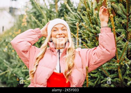 Eine niedliche Blondine wählt einen Weihnachtsbaum für das neue Jahr. Eine Frau in einem Hut und einer rosa Jacke lächelt bei schneebedecktem Wetter auf die Kamera. Winterferien und n Stockfoto