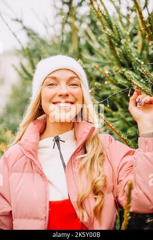 Eine niedliche Blondine wählt einen Weihnachtsbaum für das neue Jahr. Eine Frau in einem Hut und einer rosa Jacke lächelt bei schneebedecktem Wetter auf die Kamera. Winterferien und n Stockfoto