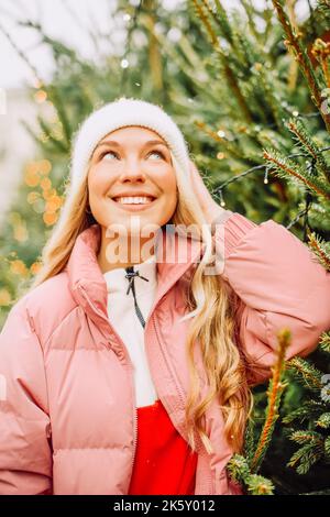 Eine niedliche Blondine wählt einen Weihnachtsbaum für das neue Jahr. Eine Frau in einem Hut und einer rosa Jacke lächelt bei schneebedecktem Wetter auf die Kamera. Winterferien und n Stockfoto