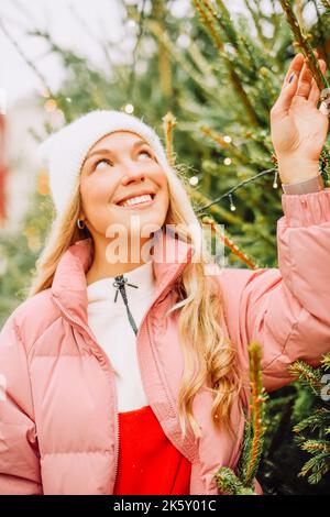 Eine niedliche Blondine wählt einen Weihnachtsbaum für das neue Jahr. Eine Frau in einem Hut und einer rosa Jacke lächelt bei schneebedecktem Wetter auf die Kamera. Winterferien und n Stockfoto