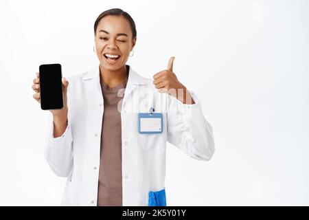 Ärztin zeigt Daumen nach oben und Smartphone-Bildschirm-App, mobile Healthcare-Anwendungsschnittstelle, stehend auf weißem Hintergrund Stockfoto