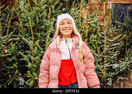 Eine niedliche Blondine wählt einen Weihnachtsbaum für das neue Jahr. Eine Frau in einem Hut und einer rosa Jacke lächelt bei schneebedecktem Wetter auf die Kamera. Winterferien und n Stockfoto