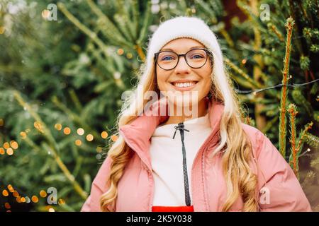 Eine niedliche Blondine wählt einen Weihnachtsbaum für das neue Jahr. Eine Frau in einem Hut und einer rosa Jacke lächelt bei schneebedecktem Wetter auf die Kamera. Winterferien und n Stockfoto