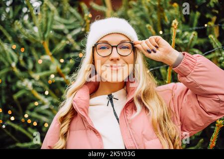 Eine niedliche Blondine mit Hut und rosa Jacke und Brille steht vor dem Hintergrund von Weihnachtsbäumen. Das Mädchen wählt einen Weihnachtsbaum, Neujahr m Stockfoto