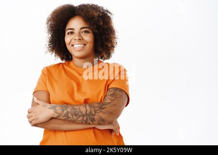 Porträt einer selbstbewussten lächelnden jungen afroamerikanischen Frau, die die Arme kreuzt, Tattoos am Arm trägt, glücklich aussieht, in orangefarbenem T-Shirt über Weiß steht Stockfoto