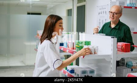 Asiatischer Apotheker hilft verschiedenen Kunden mit Krankheiten, indem er pharmazeutische Produkte aus Apotheken-Regalen gibt. Mitarbeiter, der Menschen medizinische Hilfe leistet und Medikamente in der Drogerie erklärt. Stockfoto