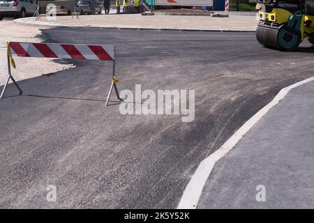 Asphalt wird von einer Schubkarre entladen. Der hintere Teil des Asphalts ist fertig gerollt. Asphaltfertiger im Hintergrund Stockfoto