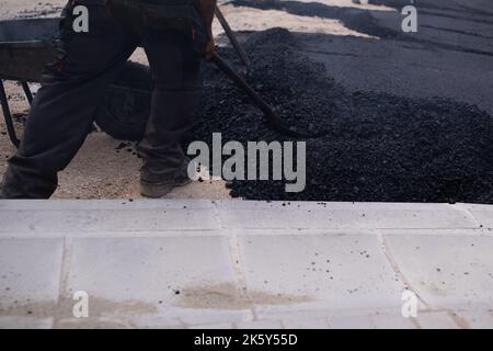 Asphalt wird von einer Schubkarre entladen. Der hintere Teil des Asphalts ist fertig gerollt. Asphaltfertiger im Hintergrund Stockfoto