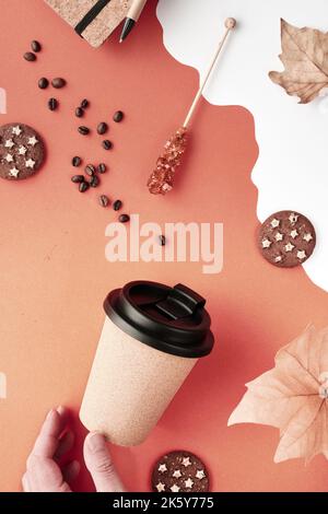 Kaffeebecher mit Naturkorkband. Kaffeetasse auf orange-weißem Herbsthintergrund mit trockenen Blättern zu gehen. Kaffeebohnen, Schokoladenkekse und Zuckerstick Stockfoto