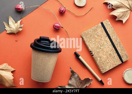 Kaffeebecher mit Naturkorkband. Kaffeebecher zu trockenen Platanenblättern und Deko-Kürbissen. Notizbuch mit passendem Korkbezug. Flach auf orange legen Stockfoto