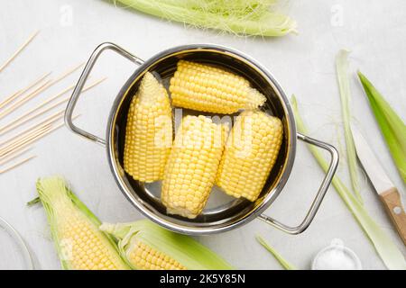 Kochtopf aus gelben süßen Maiskolben, vorbereitet zum Kochen, rohe Maiskolben, Holzspieße, Messer und Salz auf Küchentisch, Draufsicht. Stockfoto