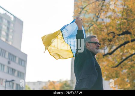 Grauhaariger, eleganter kaukasischer Mann mit blau-gelber ukrainischer Flagge in der Luft und zu Fuß im Freien. Protest. Menschen gegen den Krieg. Hochwertige Fotos Stockfoto