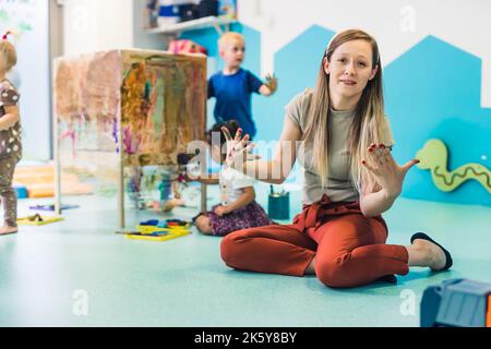 Kaukasische Lehrerin, die mit ihren gemalten Händen auf dem Boden sitzt und die Kamera anschaut, Kinder malen mit ihren Händen im Hintergrund, Kindergartenkonzept. Hochwertige Fotos Stockfoto